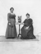 Dvě Evropanky v ateliéru u malého stolku – jedna stojí, druhá (těhotná) sedí