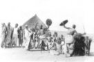 Dva muži tančící se štíty a biči před skupinou žen a dětí
