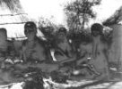 Skupina žen s dětmi sedící před chýší u ohniště s varnými kameny
