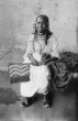 Žena v bílých šatech sedící na lehátku, v ruce drží vějíř
