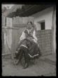 děvče sedící na dvoře, v polosvátečním kroji, hladký účes s vlasy staženými do týlu, zdobené stuhami