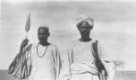Poprsí dvou mužů stojících vedle sebe, jeden má turban, druhý čapku