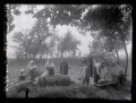 Rodina cikánů před hliněnou chatou; před chatou vyskládány proutěné koše, muž porcující zabité sele