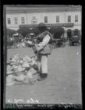 Ovčák na trhu stojí u kloboučnického zboží, v pozadí patrové měšťanské domy, lemující náměstí.