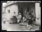 Skupina žen a dětí v tradičních krojích sedících před domem.